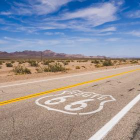 Historic Route 66 road marking along the desert, California portion of the route, with a bright blue sky and wispy white clouds above the rugged mountain horizon.