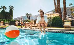 A man cannonballs into a sparkling blue pool. He is wearing swim trunks and an unbuttoned tropical shirt. Multiple palm trees are in the background. A colorful beach ball floats on the water. Other talent relax on beach furniture behind him.