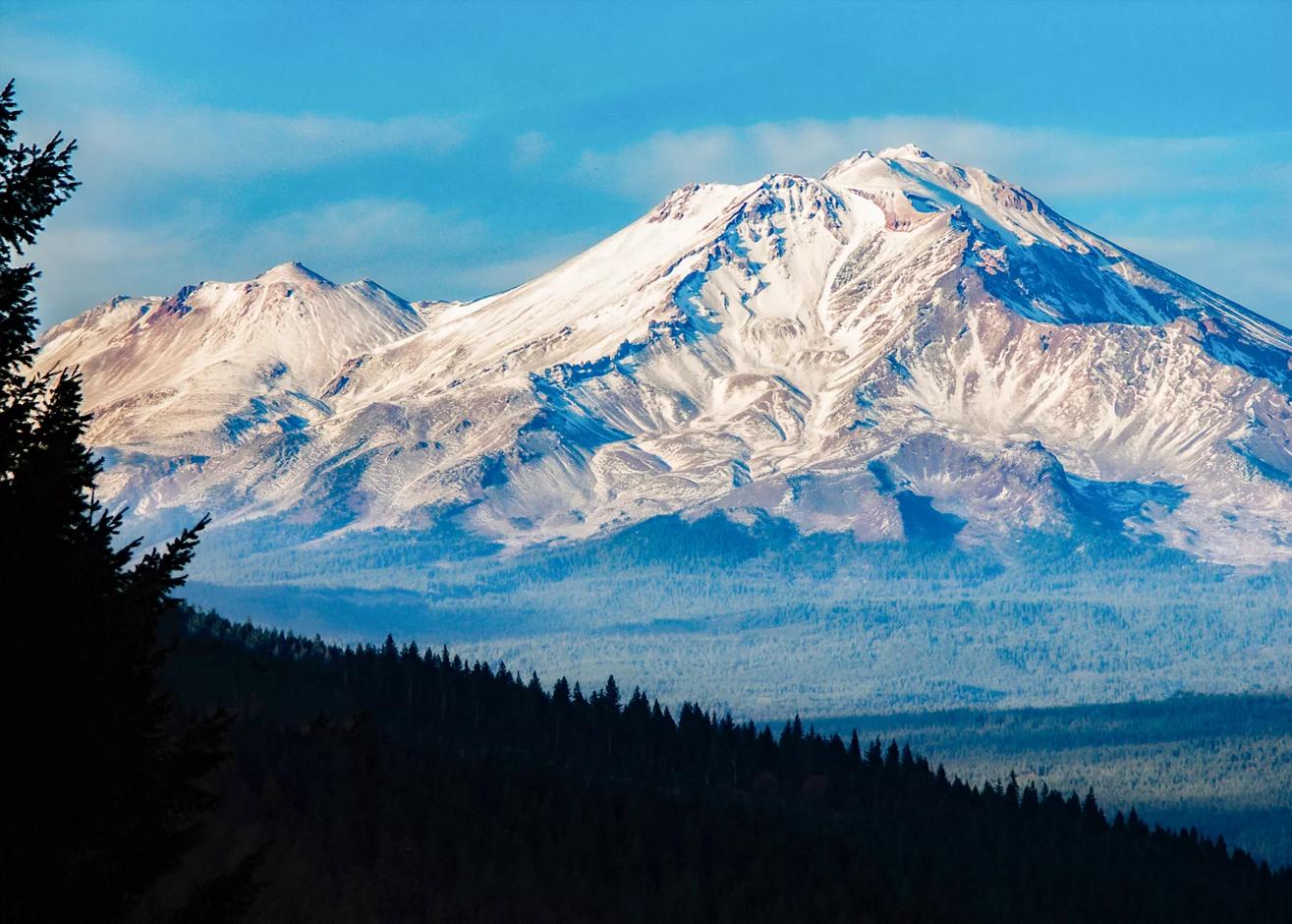 The iconic Mount Shasta is fully displayed. The image is taken from Castle Crags State Park (20022 Castle Creek Road, Castella California) The image is taken in the early morning light and is framed by evergreen trees in silhouette.  There is copy space at the bottom of the image